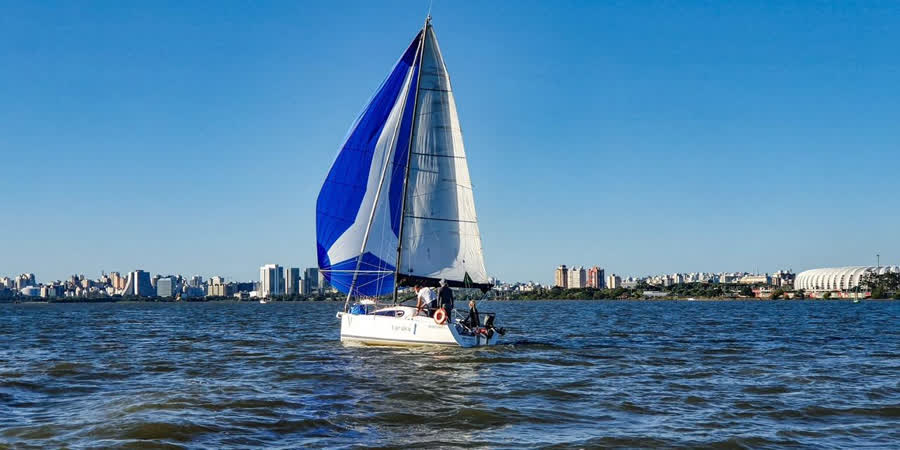 Veleiro esportivo parceiro com vela azul e branca navegando no Guaíba, com o centro de Porto Alegre ao fundo